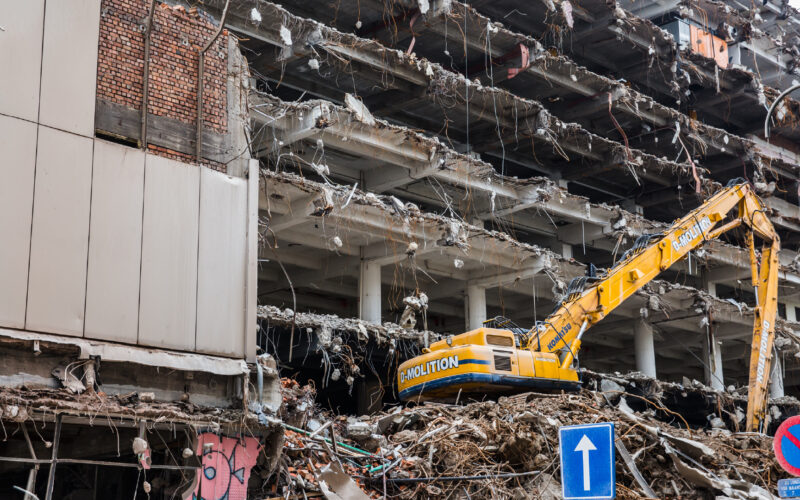 Sloopterrein met een grote gele graafmachine die actief werkt aan het puin van een gedeeltelijk gesloopt gebouw.
