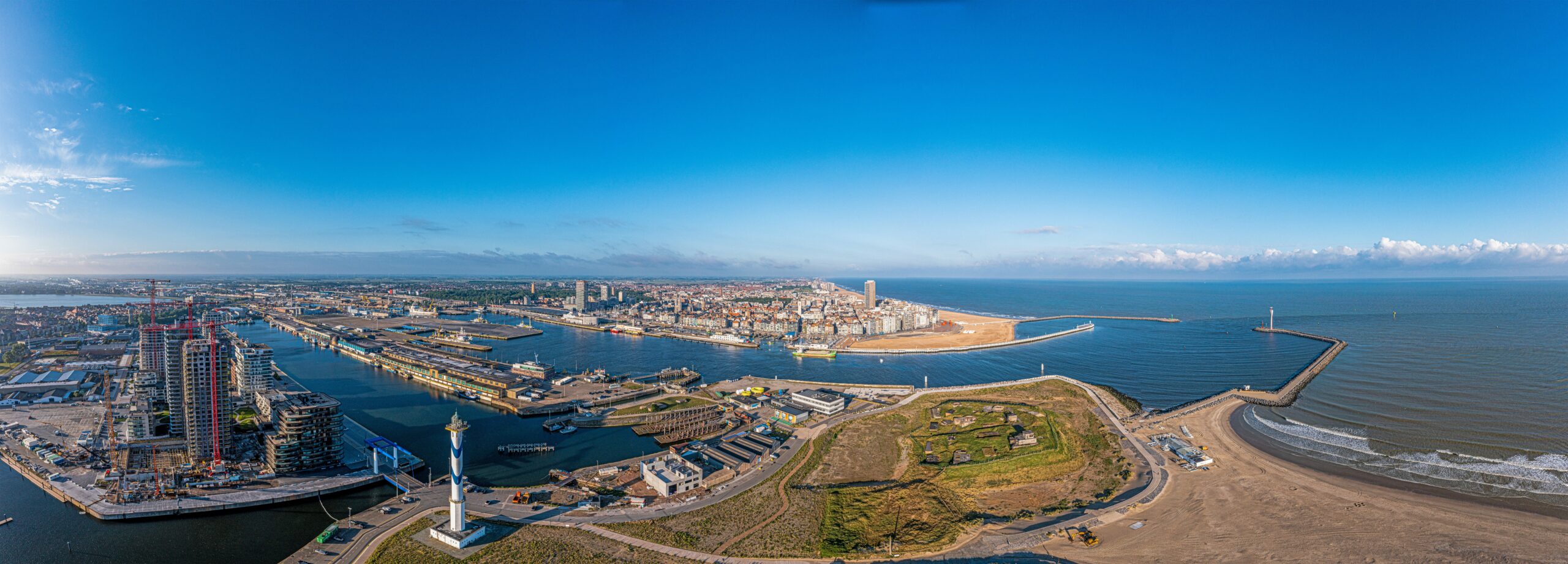 Panoramafoto van een kuststad met een haven, stranden en stedelijke gebieden onder een heldere blauwe lucht.